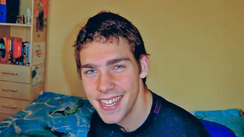 Portrait d’un jeune homme souriant, assis dans une chambre.